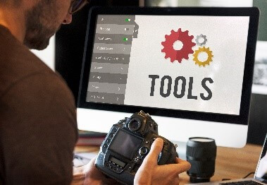 A person holding a camera while sitting at a desk with a computer displaying a screen with various tools and gears.