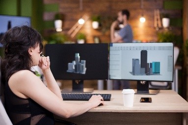 A person sitting at a desk with two computer screens, analyzing 3D architectural models, with a coffee cup and smartphone on the desk.