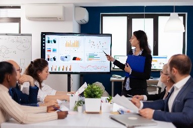 A businesswoman presenting data and graphs on a large screen to a group of colleagues in a meeting room.