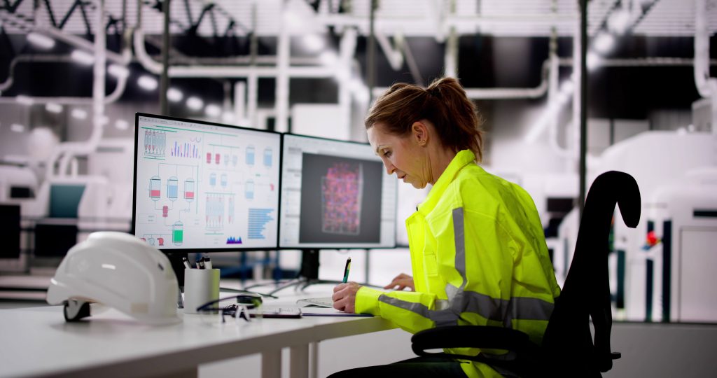 A female engineer wearing a high-visibility jacket sits at a desk in an industrial control room, analyzing data on multiple monitors displaying detailed schematics and graphs.